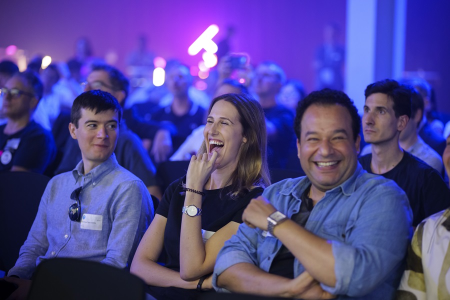 Three people sit amongst a crowd. The woman in the middle is wearing a black shirt and is laughing with her hand up to her face, her smile is infectious. The on the right is wearing a blue top and is looking at the camera with a big grin and the man to her left is looking over at them both smiling. It looks as though the presentation they are watching has made them all laugh.