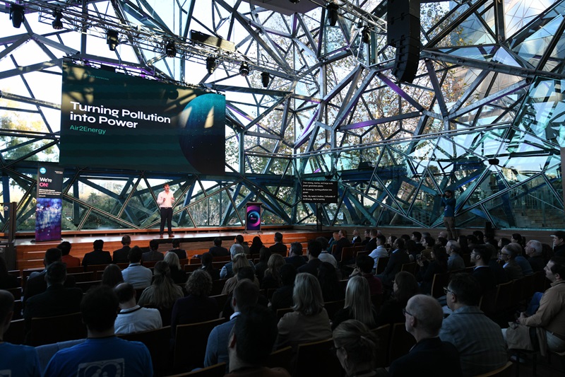 Presenter on stage speaking to a seated audience at an event venue with a geometric glass facade. A large screen behind the presenter displays the words “Turning Pollution into Power” and “Air2Energy”.