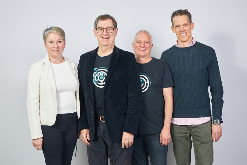 A woman and 3 men are standing close together in front of a white background, smiling at the camera. 