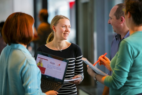 A group of three females and a male stand having a conversation. One blond woman stands facing the camera with an open and engaging expression on her face. The other people stand with their backs to the camera.