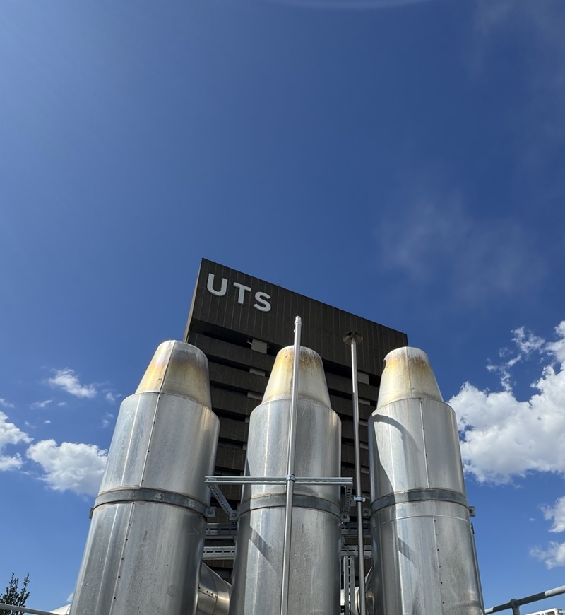 Three large cylindrical metal units installed outdoors in front of a UTS building. The photo is taken from below against a blue sky with scattered clouds.
