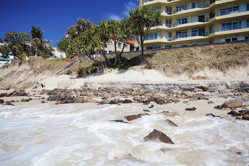 A beach is eroded in the foreground with apartment building in the background