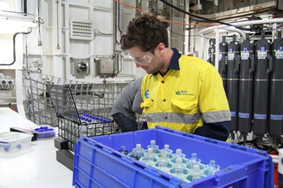 A man looks over a series of bottles in a blue tub. The CTD profiler aboard Investigator is visible in the background.