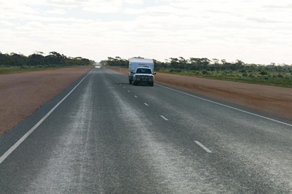 Long stretch of Australian road with a car towing a caravan with road lines in foreground and red dirt either side of road