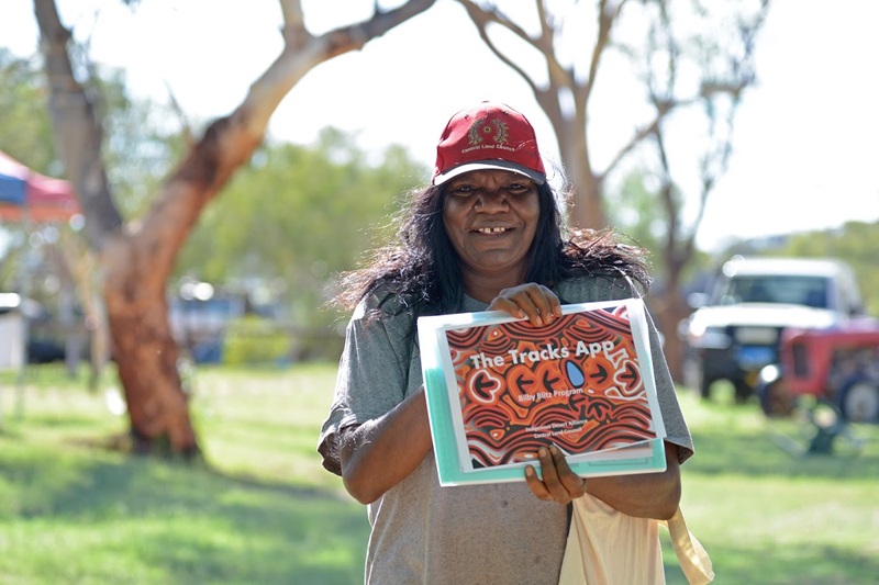 Picture of an Indigenous woman wearing a Central Land Council hat, smiling for the camera and holding up a poster that reads "The Tracks App"