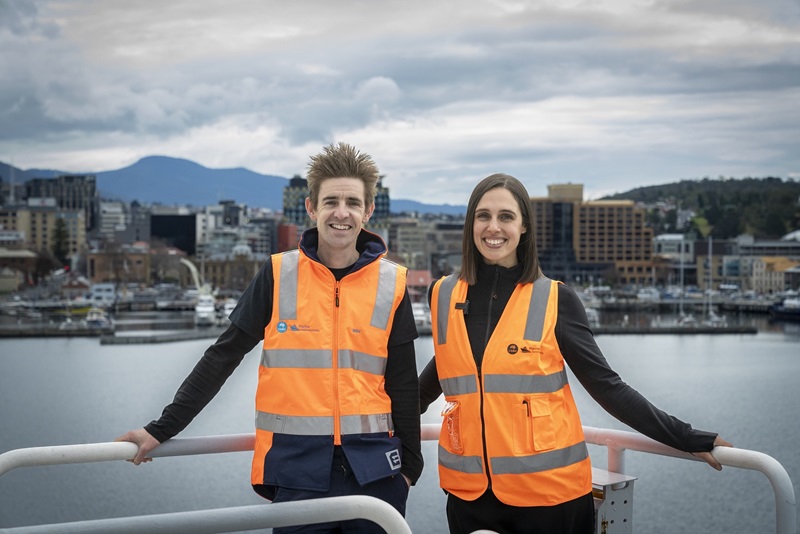 Man and woman in hi-vis vest standing on deck of ship with Hobart in background