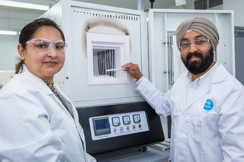 A woman and man dressed in white coats in a laboratory setting stand by a furnace.