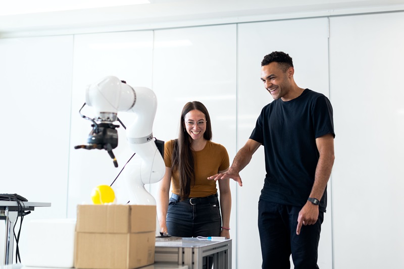 Two smiling people gesture towards a robot with a mechanism for lifting soft objects 
