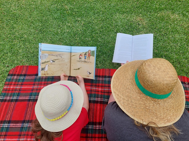 Top-down view of two people lying on a blanket on the grass and reading a book.