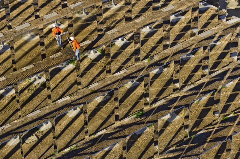 Solar field with reflective panels on dirt ground with man in HI-vis and hard hat cleaning the panel.