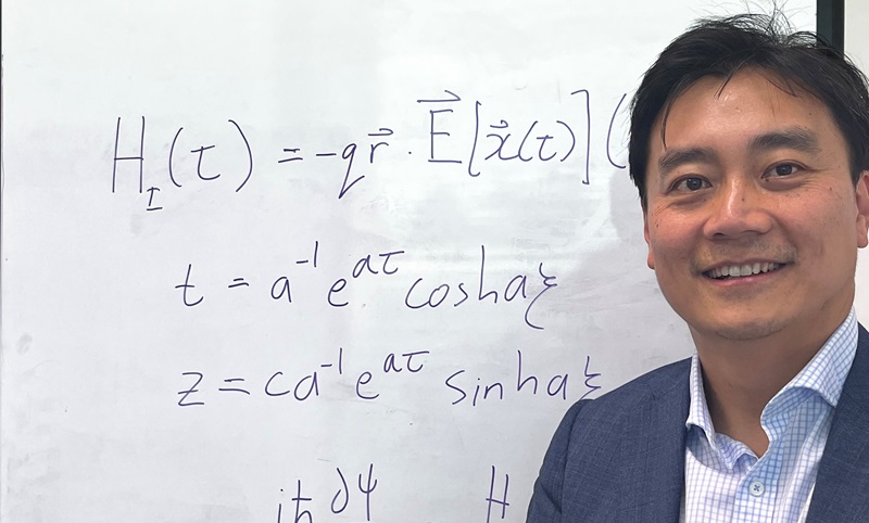 Close up shot of man wearing a blue open shirt and a suit jacket standing in front of a whiteboard with science/maths equations on it. 