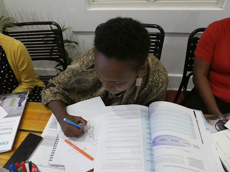 A woman with short cropped hair and a patterned shirt leans over a notebook with a pen