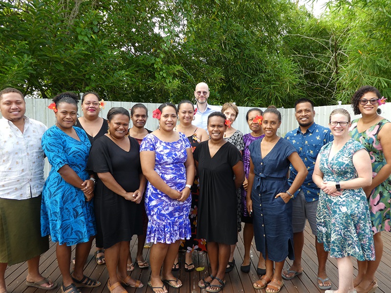 A group of 15 are smiling at the camera. Most people are wearing flip-flops and many of the women have a flower pinned in their hair. 