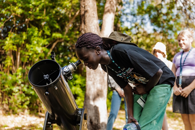 Young woman bending over to look at a large telescope in an outdoor setting. 