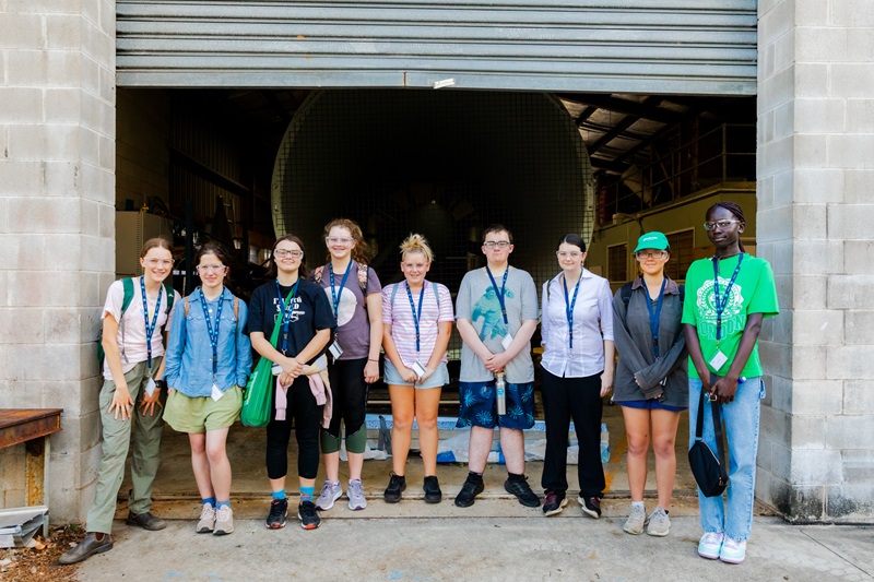 A group of young men and women, wearing lanyards, stand at the entrance to a large structure/warehouse. 