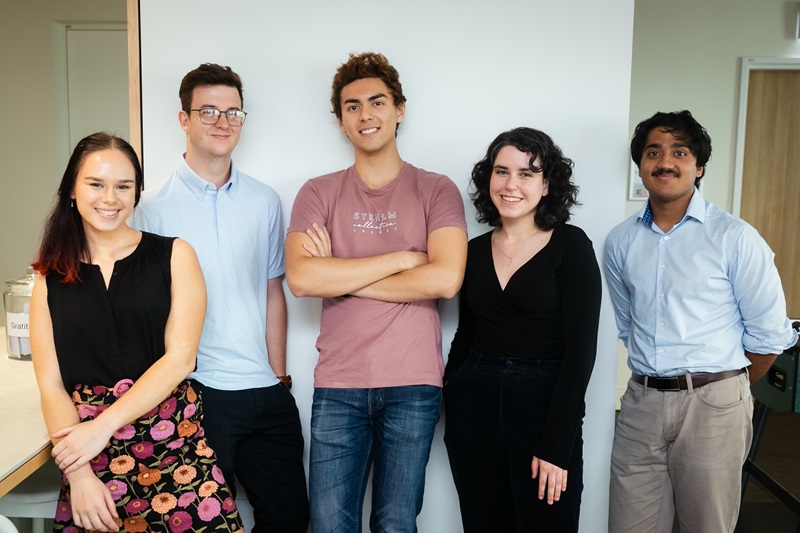 Two women and three men, all aged in their 20s, stand in front of a white wall