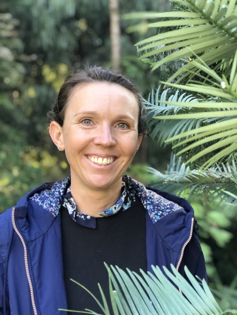 Head and shoulders of a smiling person standing beside a Wollemi Pine growing in a botanical garden.