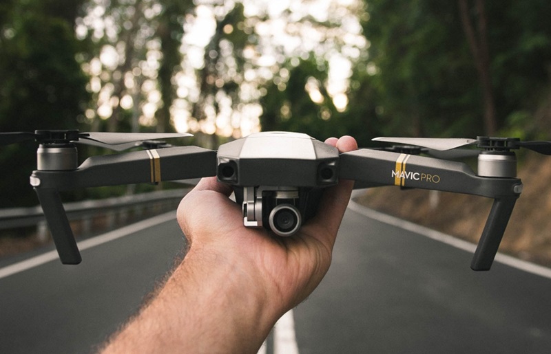 A person's hand holding a black drone. in the background is a tree lined road