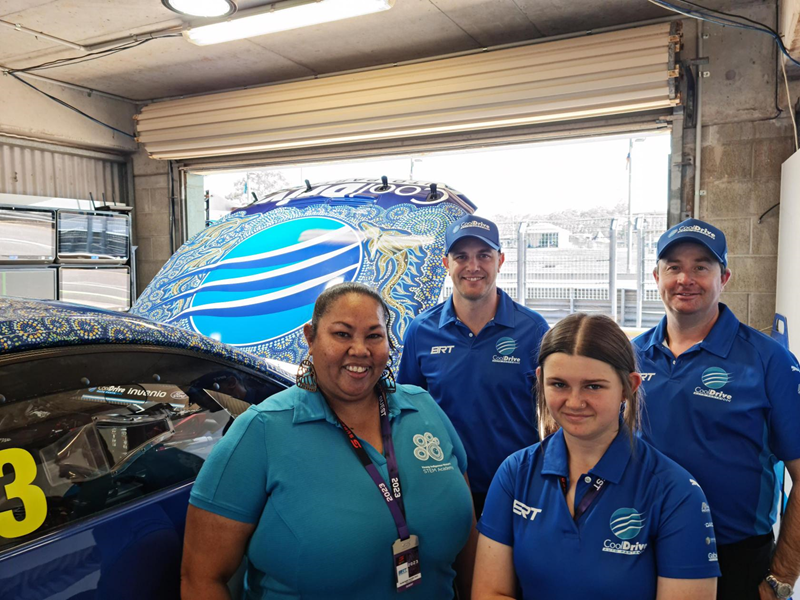 Four people in blue shirts smile at the camera. They are standing next to a racecar adorned with beautiful Indigenous art.