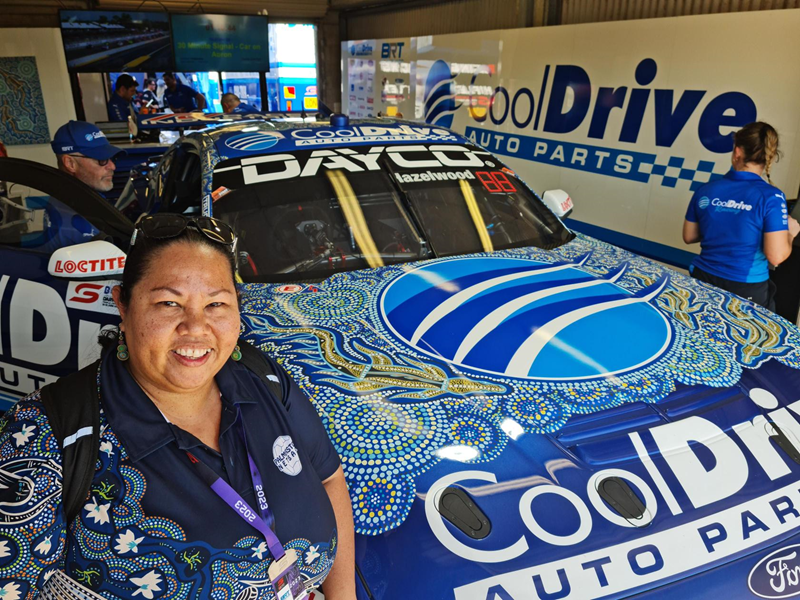 Indigenous woman stands in front of a blue racecar adorned with her beautiful Indigenous painting.