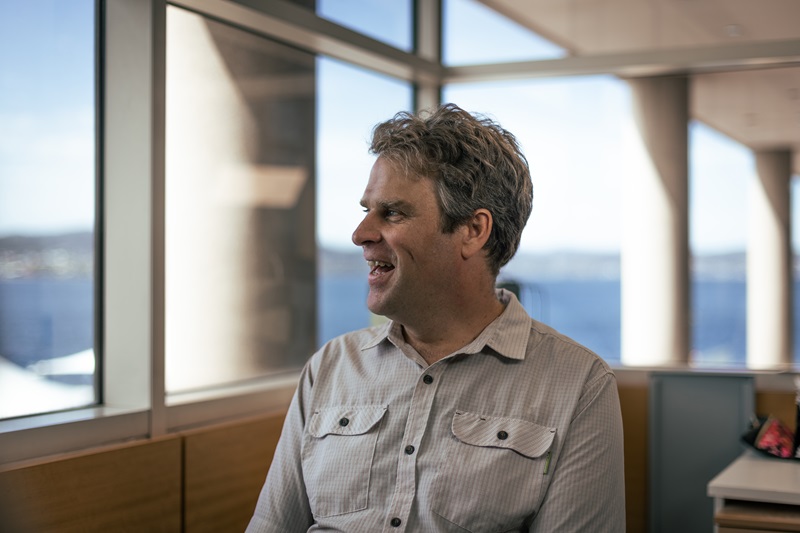 Man sits in a chair in front of some windows. He is smiling and looking to his right.