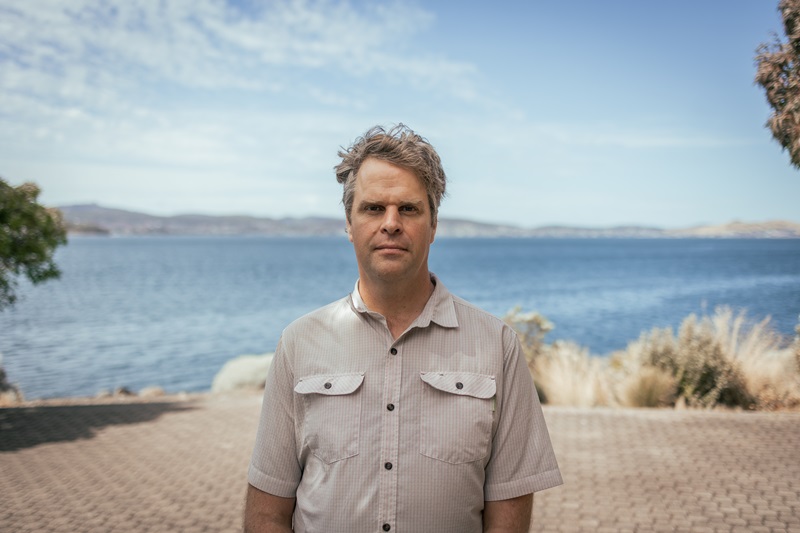 Man is standing on a path in front of a body of water. He is looking at the camera with a serious face.