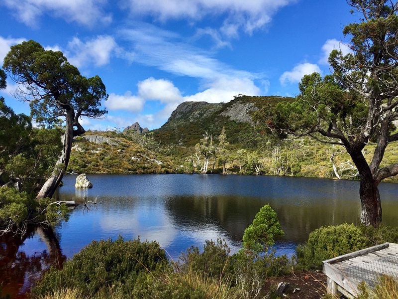 A lake in front of a mountain, surrounded by bushland, with blue sky and clouds.