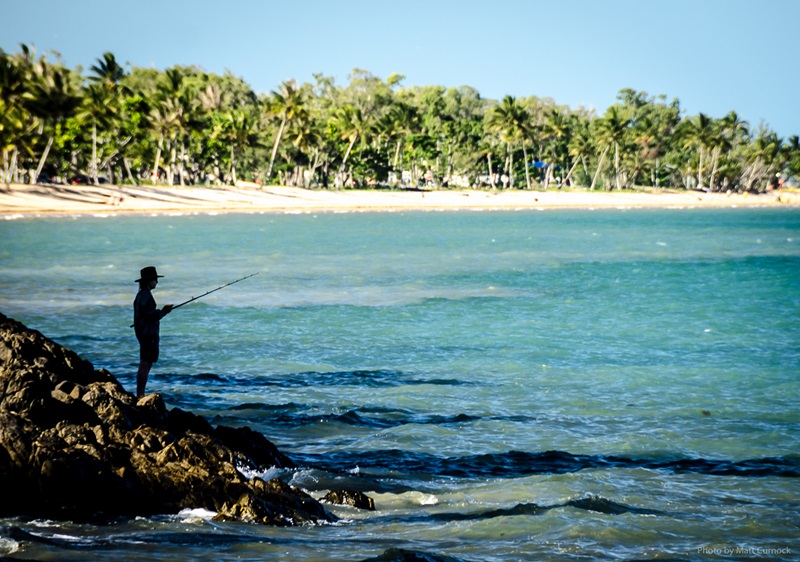 A lone fisherman with a rod fishing, the beach and shore can be seen in the background