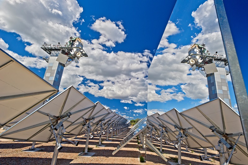 A field of solar panels are arrayed toward a tall tower against a sky of blue with a few fluffy clouds. The tower is mirrored in the surface of a panel to the right of the image. 