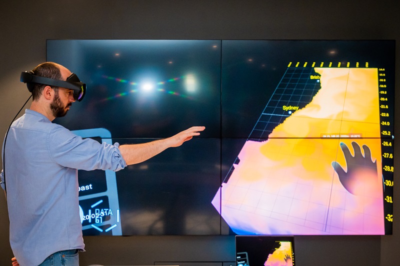 A person wearing an augmented reality headset interacts with a data visualisation of the ocean seen behind them 