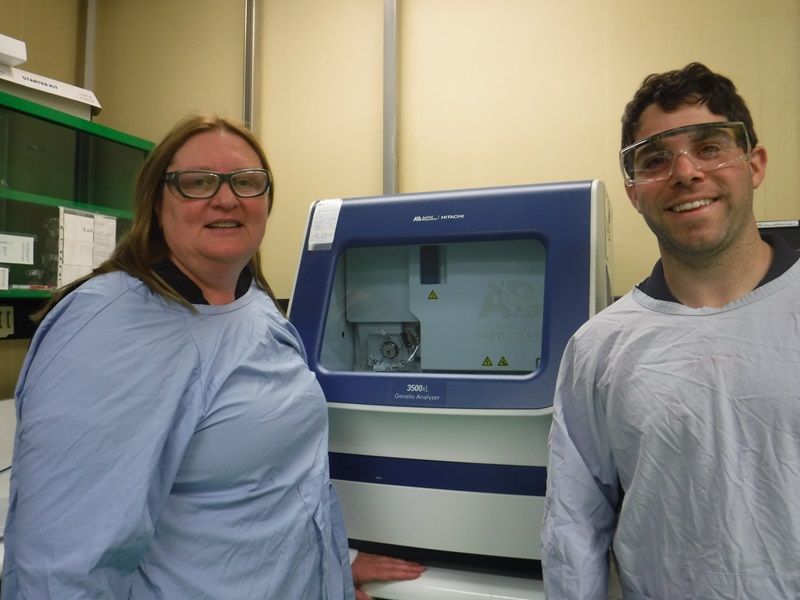 A woman and man wearing blue labcoats and safety glasses are standing in front of a piece of laboratory equipment.