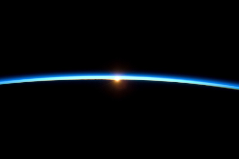 The dark sphere of the Earth is edged in a thin blue line of atmosphere, lit from behind by the rising Sun. 