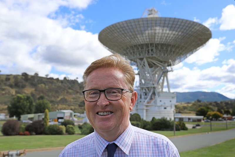 A man in glasses poses before an enormous dish antenna on a sunny day. 