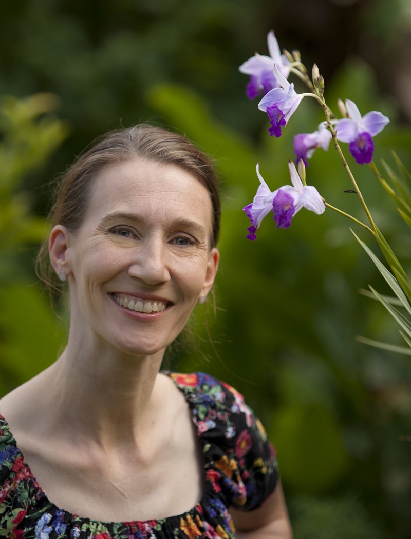 Portrait of a woman looking at the camera with a purple and white orchid in the foreground.