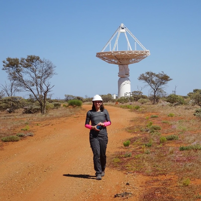 A photo of a woman standing on red soil with a large radio telescope in the background.