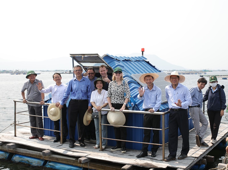 A group of people stand on a floating boardwalk. They stand in front of a shed structure. They are smiling at the camera, some of the people are giving a thumbs up sign.