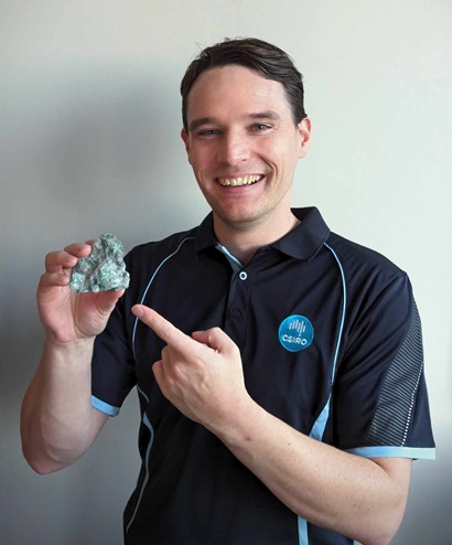 Matt Shaw in a CSIRO polo shirt smiling and holding a fist sized piece of green rock