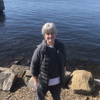 A woman with short grey hair and a relaxed smile stands in front of a body of water. 