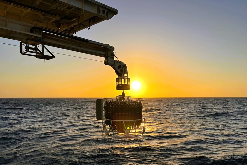A piece of scientific equipment hanging from a boom above the ocean with the sun rising above the horizon in the background.