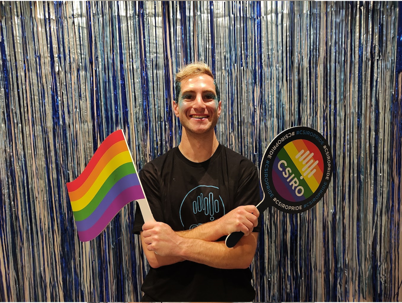 Mark holds a rainbow Pride flag, and a paddle showing the CSIRO logo on a rainbow background. Mark is smiling with joy.