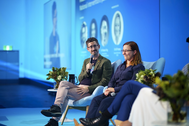 A man and a woman on a stage in conversation at a conference.