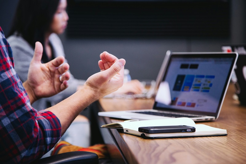 A person sitting in a front of a laptop makes a gesture while speaking at a meeting. 