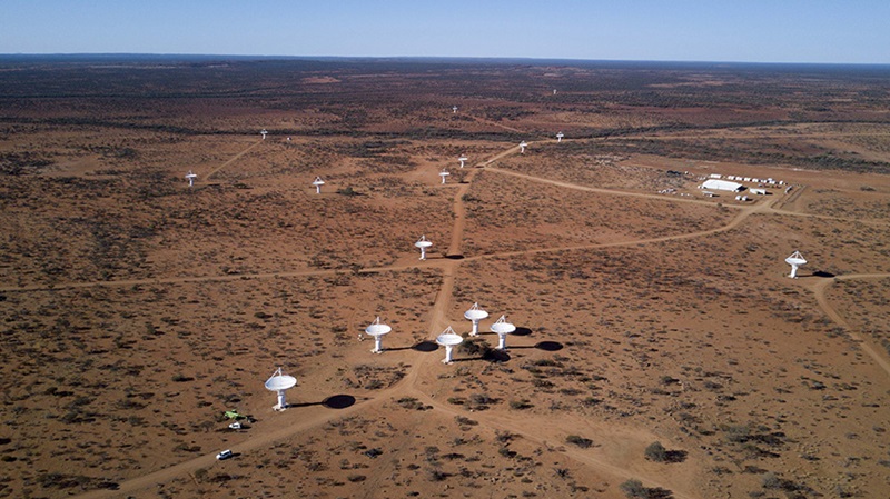 Many of the tall white dish-shaped antennas of CSIRO's ASKAP radio telescope are seen from above against the red earth of Wajarri Country. 