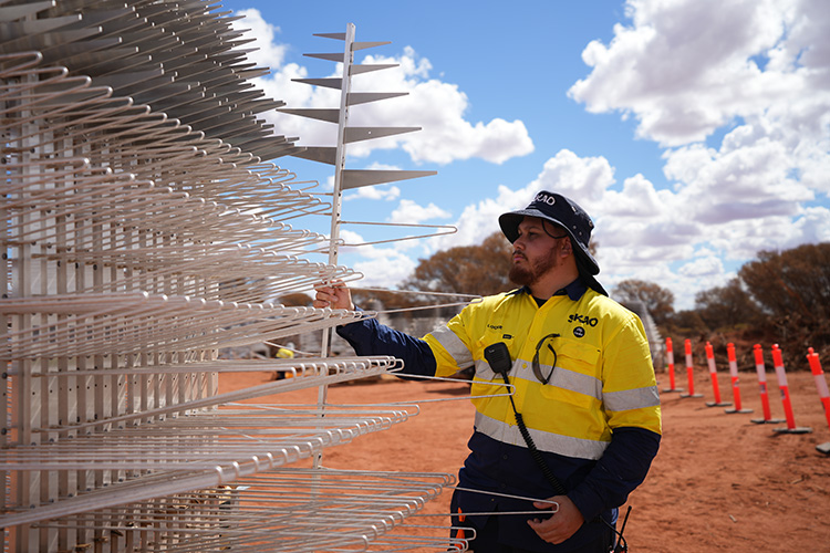 A Wajarri man in a yellow high-vis work shirt marked with the SKA Observatory and CSIRO logos and his name 'Lockie' picks up one of a stack of large silver triangular-shaped SKA-Low antenna pieces. 