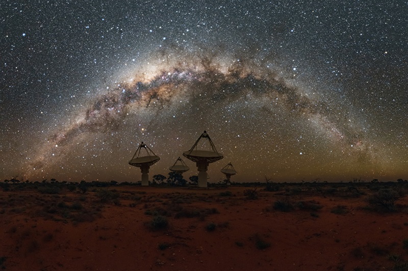 Four tall dishes of CSIRO's ASKAP radio telescope stand shadowed in the centre of the image with the dark red earth in the foreground and the brilliant arc of the Milky Way above. 