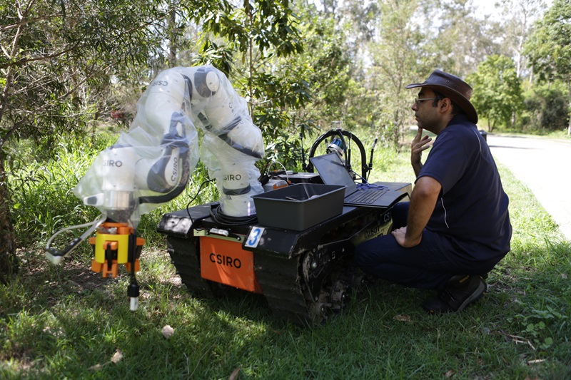 A researcher examines a tracked robot with a large claw on the front in a green field. 