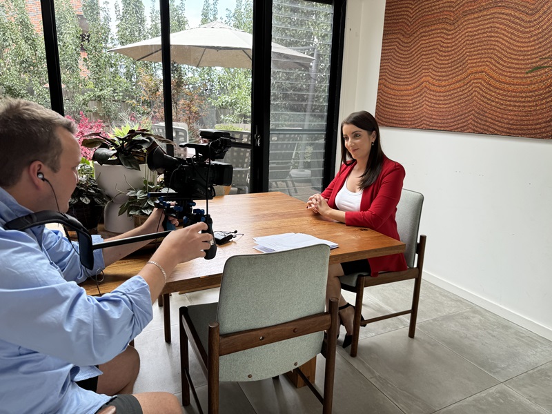 Woman sitting at table being filmed by videographer. 