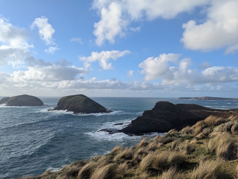 Tussocked edge of mainland on the right with islands dotted across the sea to the left under a blue sky with clouds.