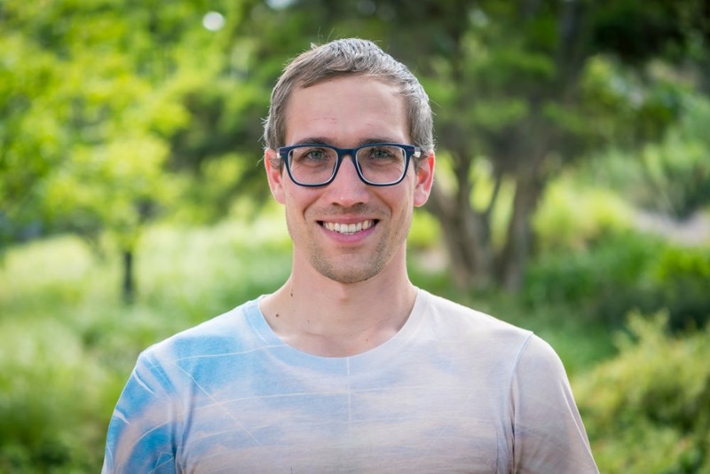 Close up of Andy wearing glasses in a t-shirt against a backdrop of green trees.