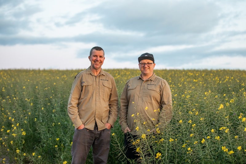 Two men wearing khaki shirts, standing in an open field of waist high grass with small yellow flowers, facing the camera.